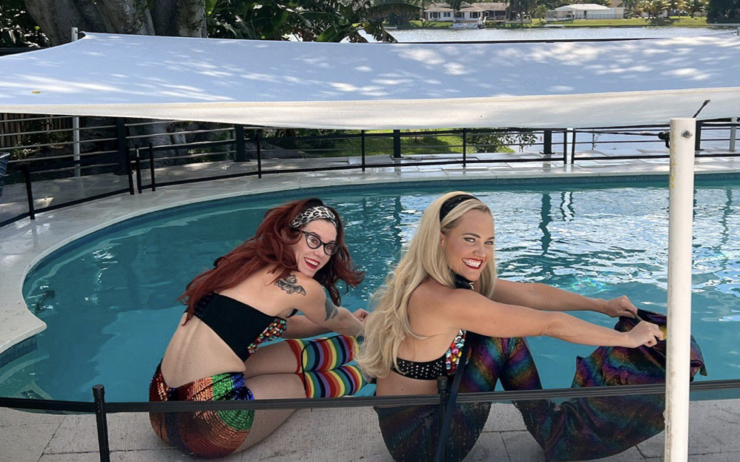 Two female swimmers, one blond, one redhead, in mermaid costumes in front of a pool.
