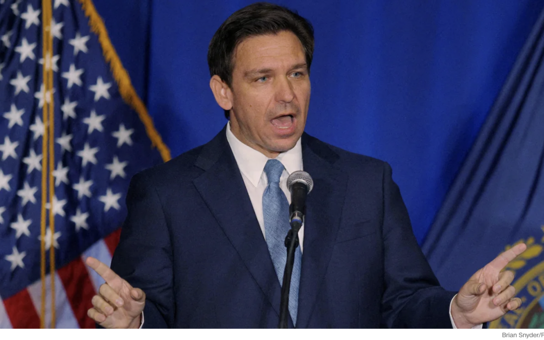 A white man with brown hair, Florida Governor Ron DeSantis in a blue suit, white shirt and blue tie against a blue backdrop and American flag.
